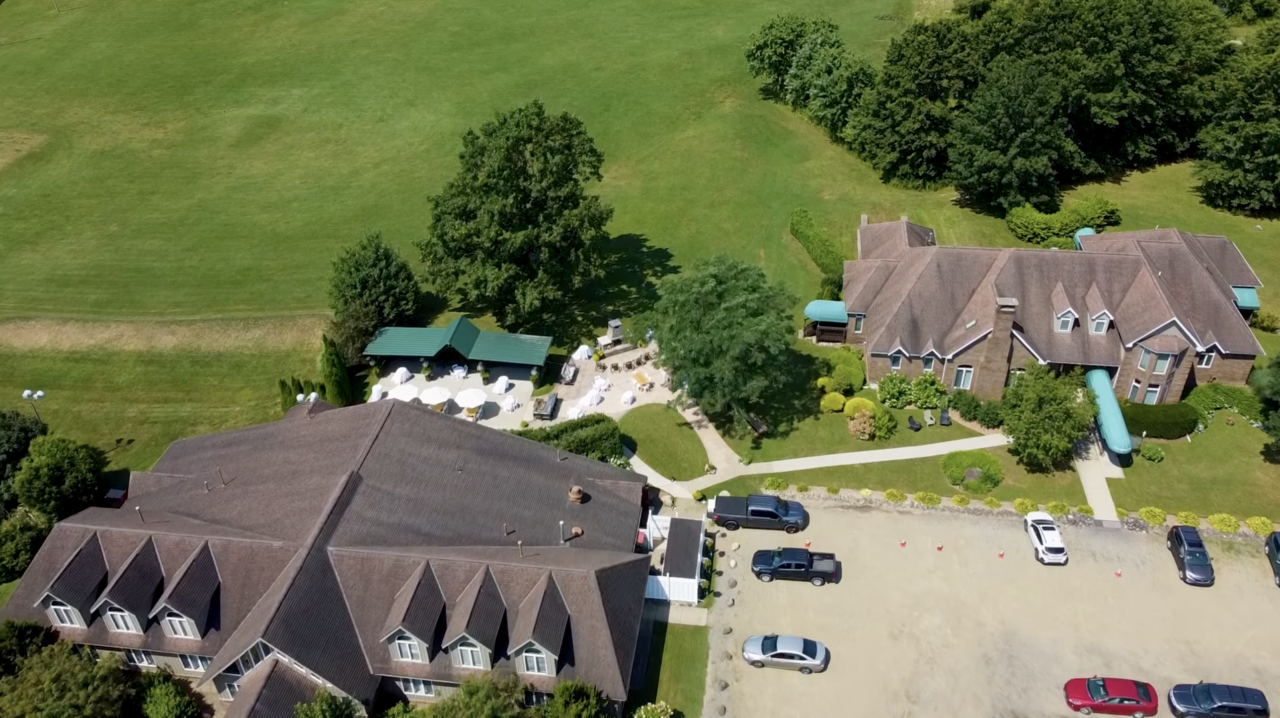 Aerial view of Nick’s Place in Edinboro showing two large buildings with brown roofs surrounded by green lawns and trees. A patio area with white umbrellas and outdoor seating sits between the buildings, while a gravel parking lot with several parked cars is visible in the foreground.
